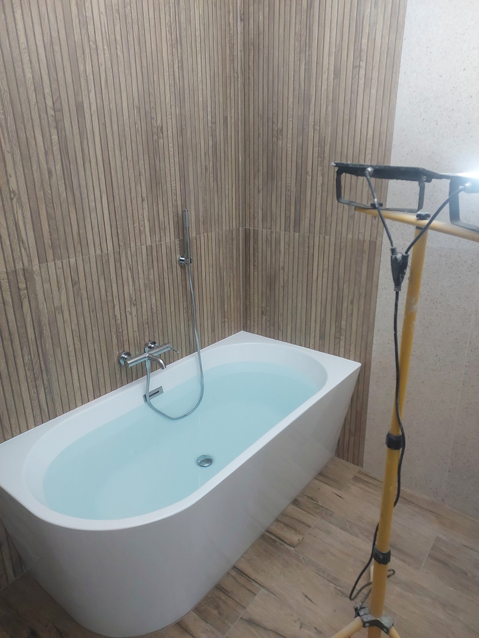 Nowoczesna wanna wypełniona wodą, z armaturą i lampą budowlaną w tle, na tle ściany z płytek imitujących drewno. Widoczny fragment podłogi z płytek.