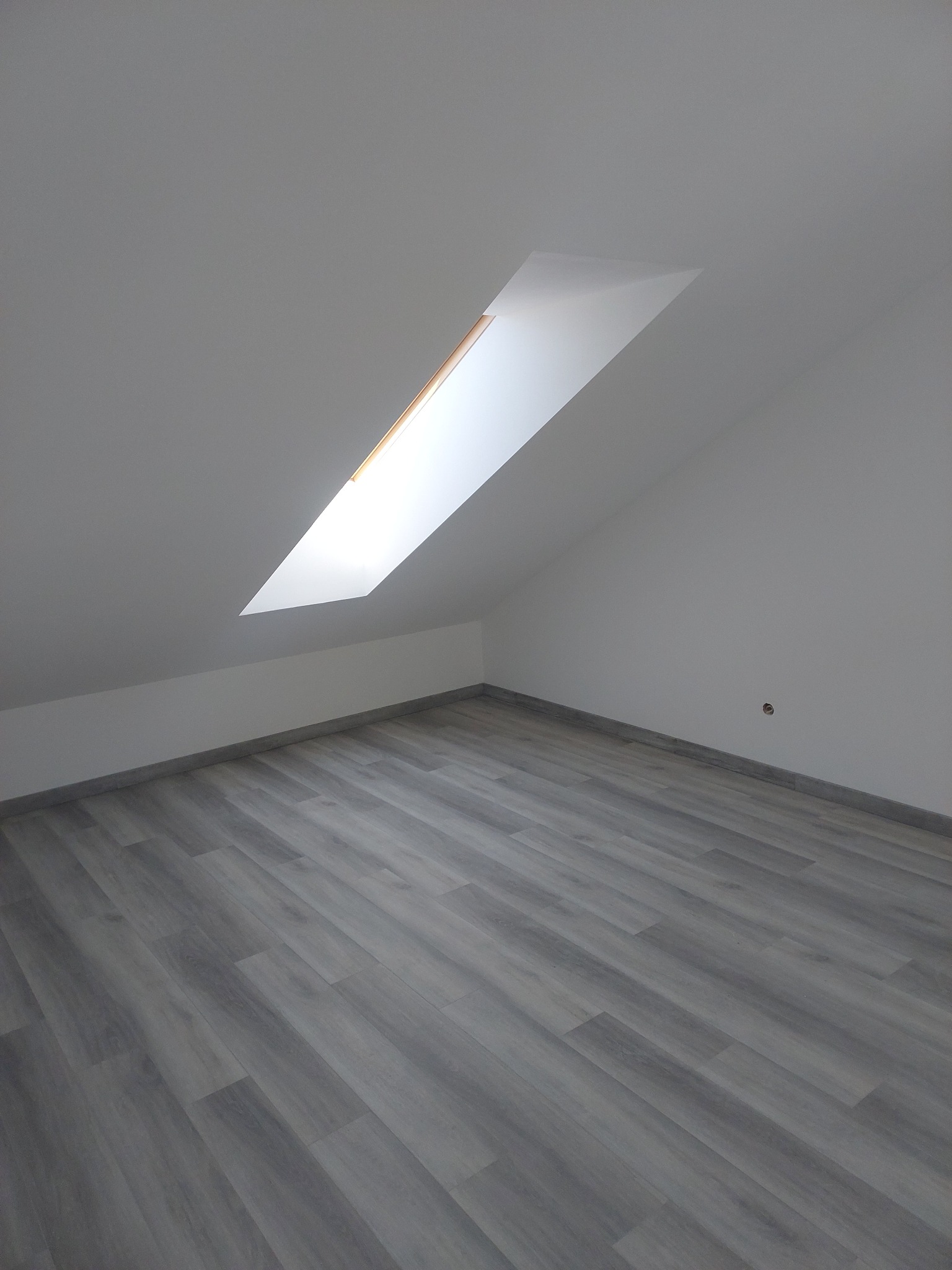 Poddasze z nową, szarą podłogą imitującą drewno. W dachu okno połaciowe wpuszczające jasne światło. Ściany pomalowane na biało, widoczny fragment gniazdka.