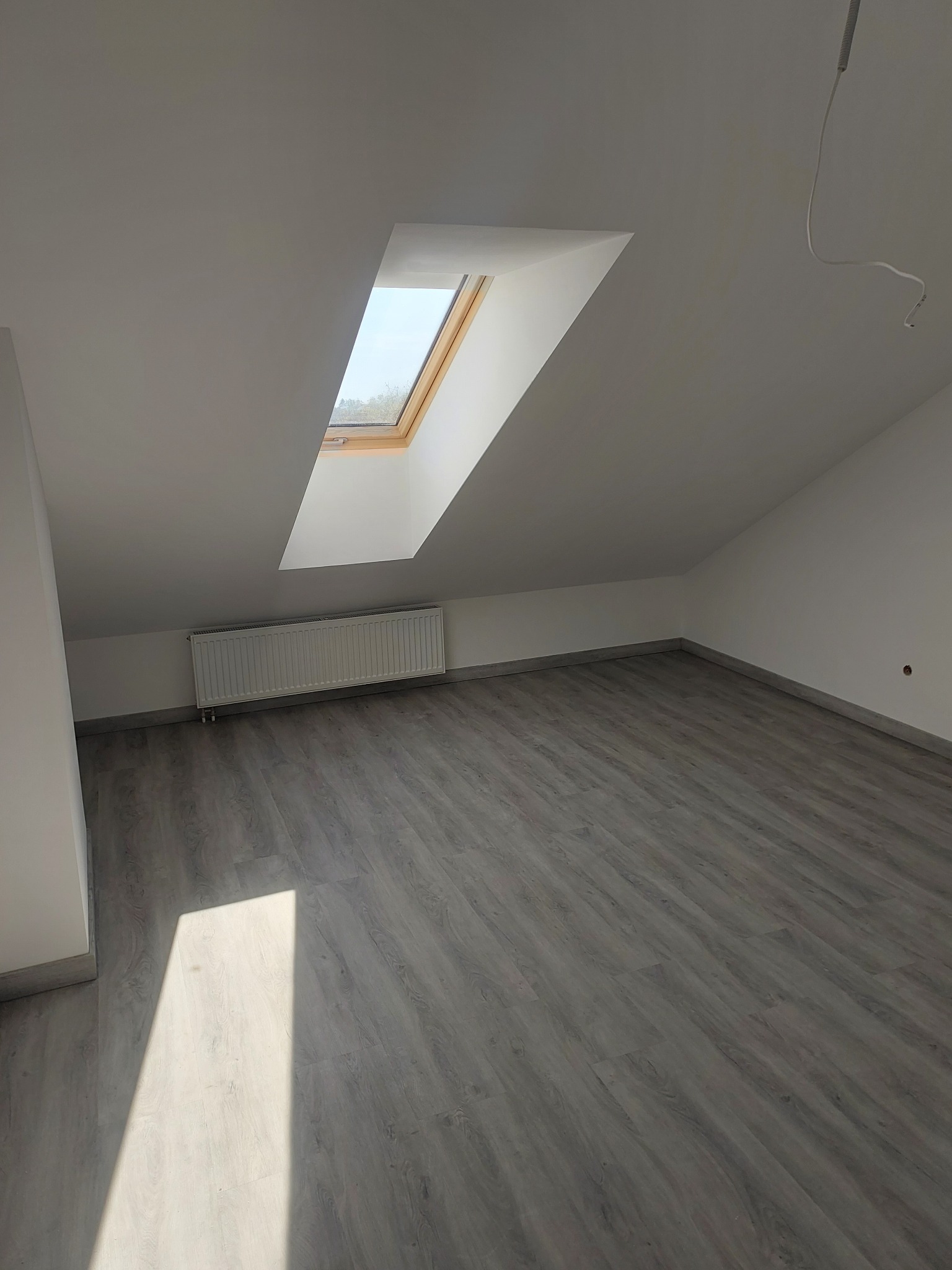 Jasne poddasze po remoncie: szara podłoga, białe ściany i okno dachowe. Grzejnik pod oknem, widoczny kabel zwisający z sufitu.