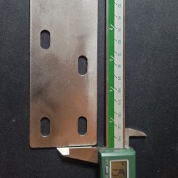 E-VOLT - Płytka metalowa z czterema otworami mierzona suwmiarką elektroniczną na czarnym tle. Widoczny wynik pomiaru na wyświetlaczu suwmiarki. Płytka o gładkiej powierzchni.