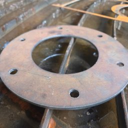 E-VOLT - Okrągły, metalowy element z otworami na mokrej, metalowej powierzchni. Wycięty detal z widocznymi śladami cięcia. Bliski plan, surowe wykończenie.