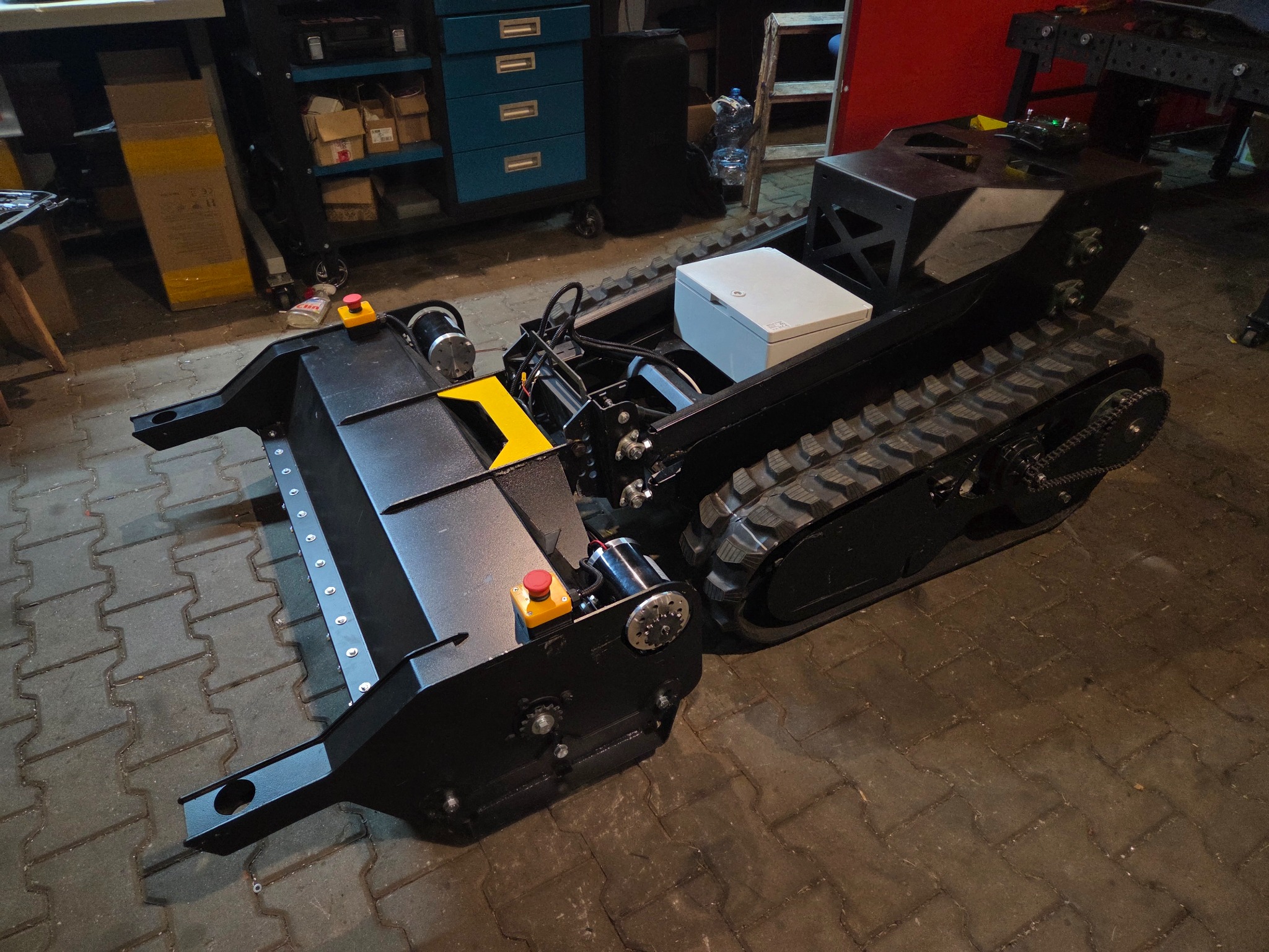 Czarny, gąsienicowy robot z żółtymi akcentami, wyposażony w lemiesz, na tle warsztatu. Widoczne detale konstrukcyjne i elementy sterujące.