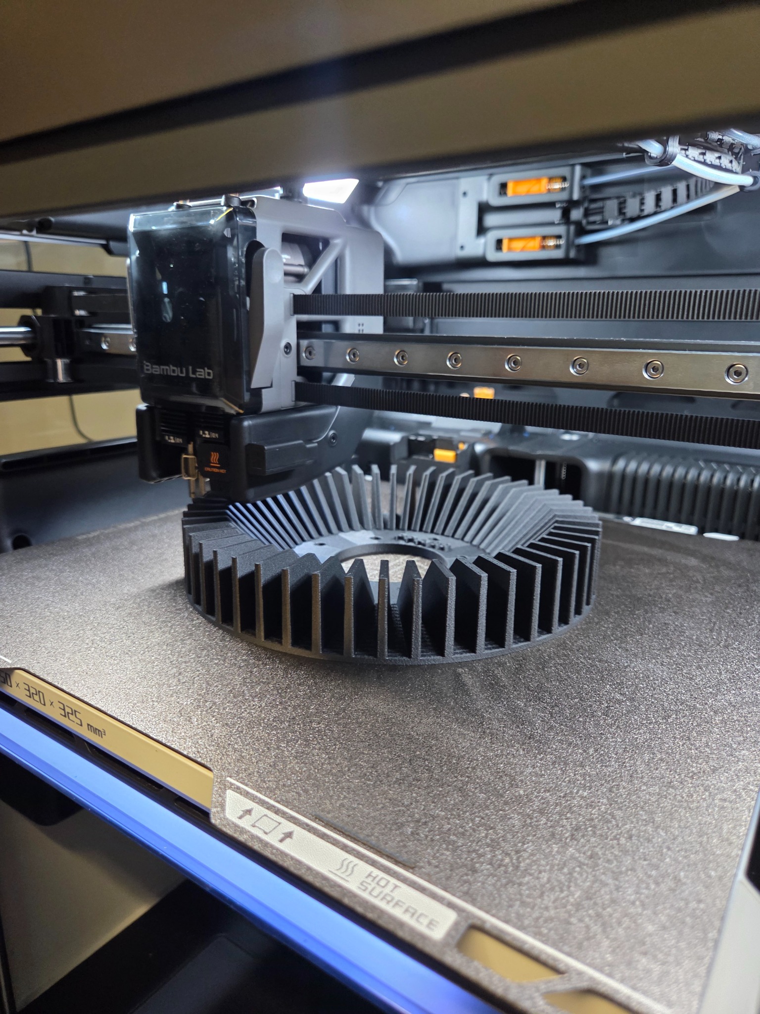 Detal z drukarki 3D Bambu Lab: czarny, okrągły element z żebrami, leżący na platformie roboczej. Widoczny mechanizm drukarki i ostrzeżenie o gorącej powierzchni.
