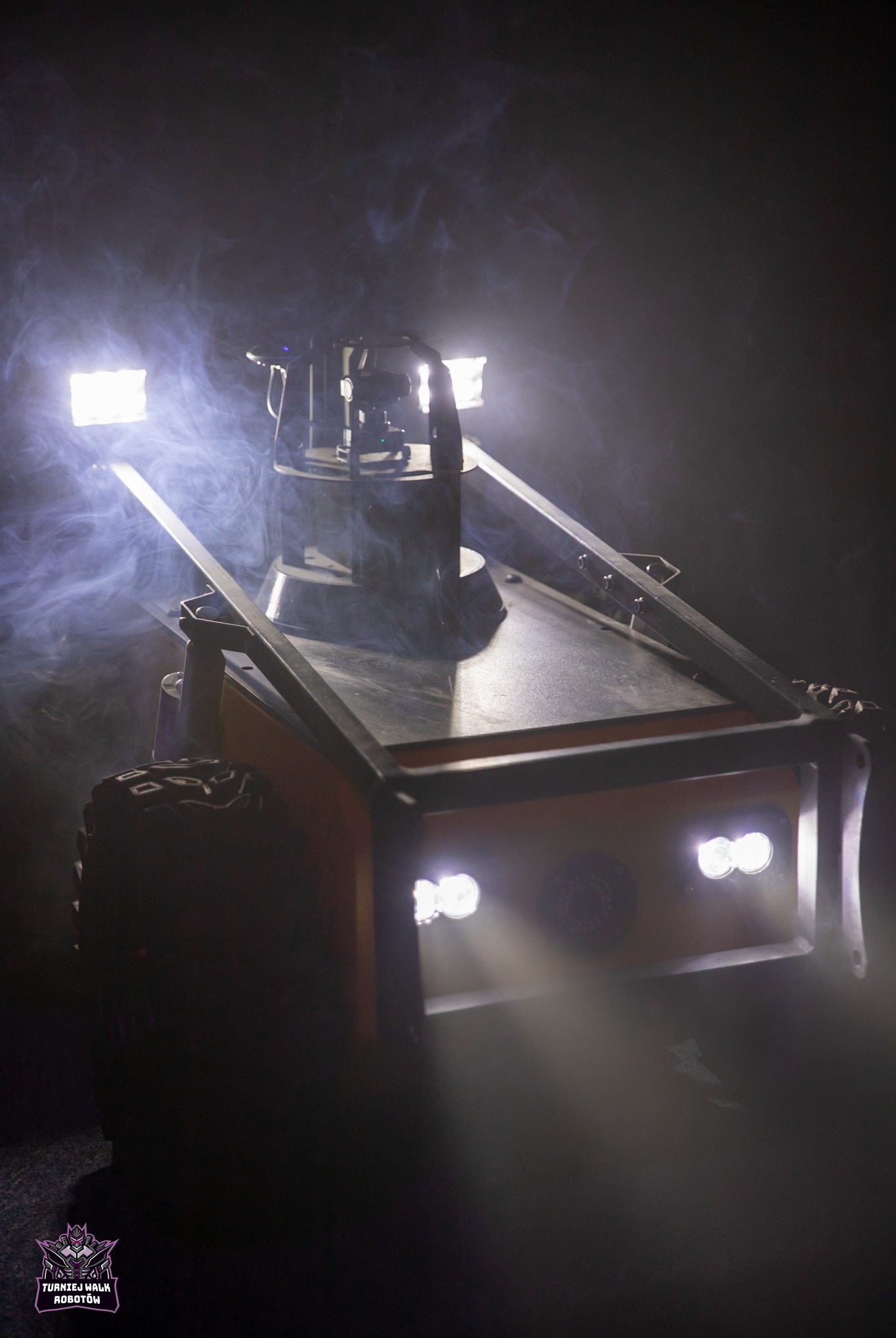 Żółta, metalowa konstrukcja z oświetleniem LED w dymie, na kołach. Widoczne detale spawania. Nietypowe ujęcie z dołu, podkreślające techniczną naturę.