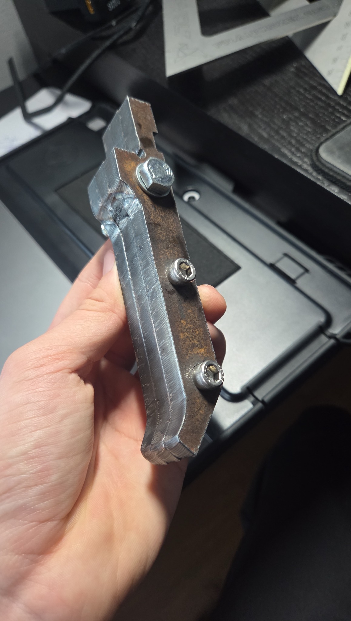Metalowy element z trzema śrubami, trzymany w dłoni, z widocznymi śladami obróbki i spawania. W tle narzędzia i powierzchnia robocza.