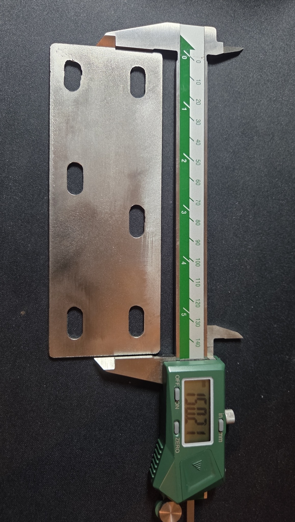 Płytka metalowa z czterema otworami mierzona suwmiarką elektroniczną na czarnym tle. Widoczny wynik pomiaru na wyświetlaczu suwmiarki. Płytka o gładkiej powierzchni.