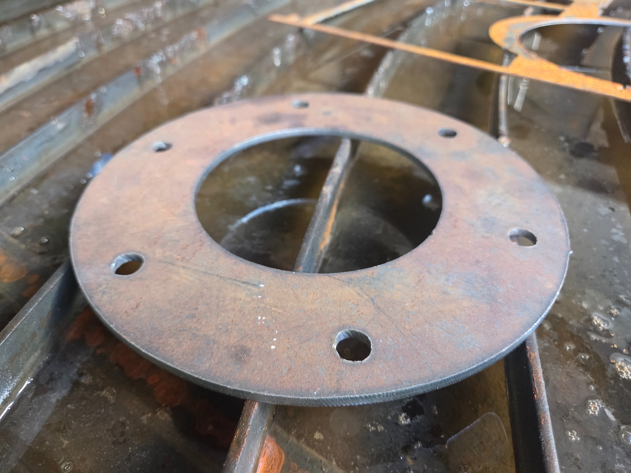 Okrągły, metalowy element z otworami na mokrej, metalowej powierzchni. Wycięty detal z widocznymi śladami cięcia. Bliski plan, surowe wykończenie.