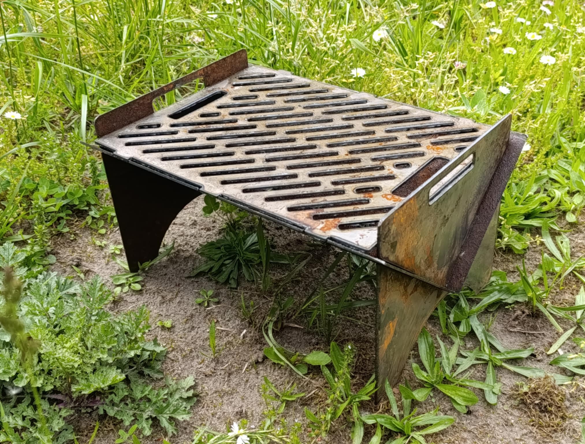 Metalowy, przenośny grill z rdzawymi elementami, ustawiony na ziemi wśród zielonej trawy i drobnych kwiatów. Widoczne spawane łączenia i ażurowa konstrukcja.