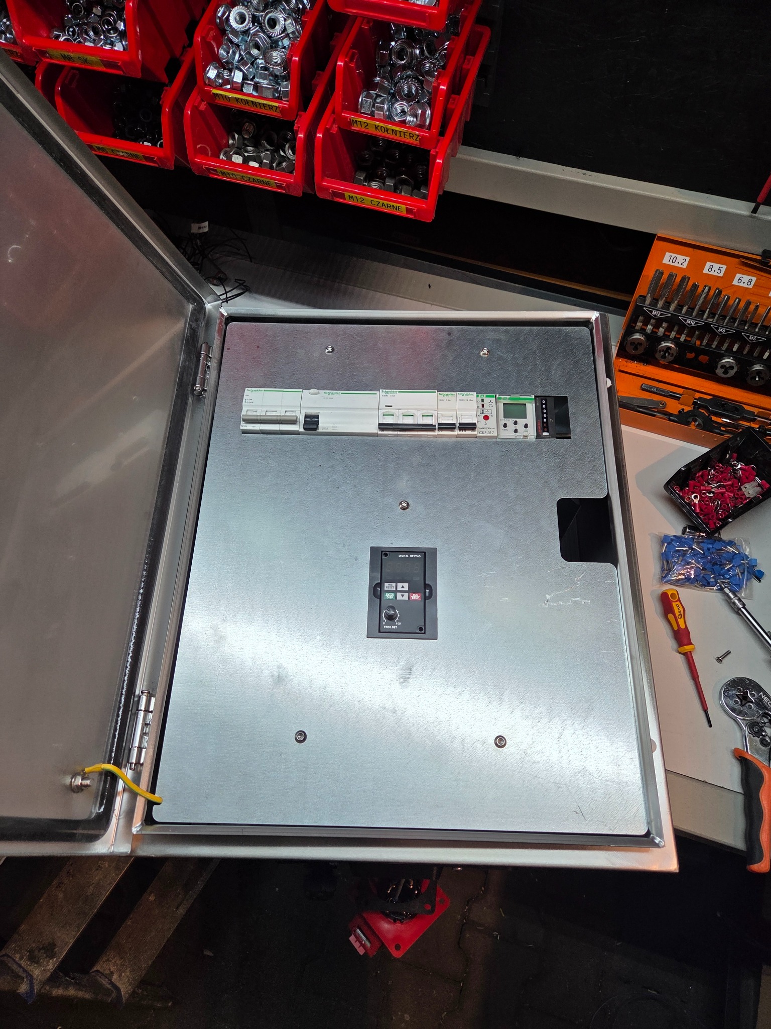 Otwarta metalowa szafa sterownicza z zamontowaną aparaturą elektryczną, widoczne narzędzia i elementy montażowe. Górny kadr z pojemnikami na śruby.