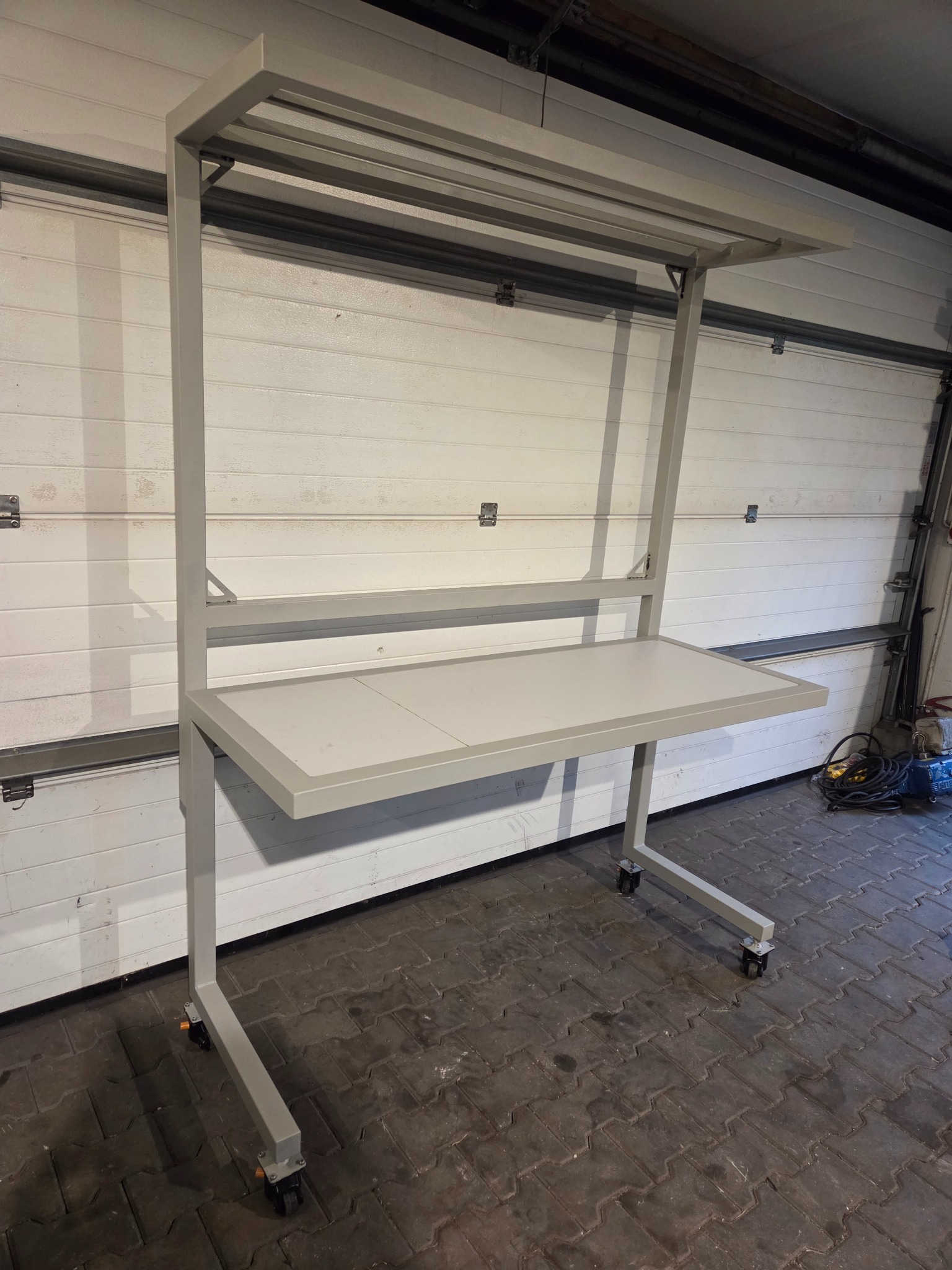 Metalowa konstrukcja na kółkach z blatem roboczym, szara, spawana, widoczna na tle białej bramy garażowej i kostki brukowej.
