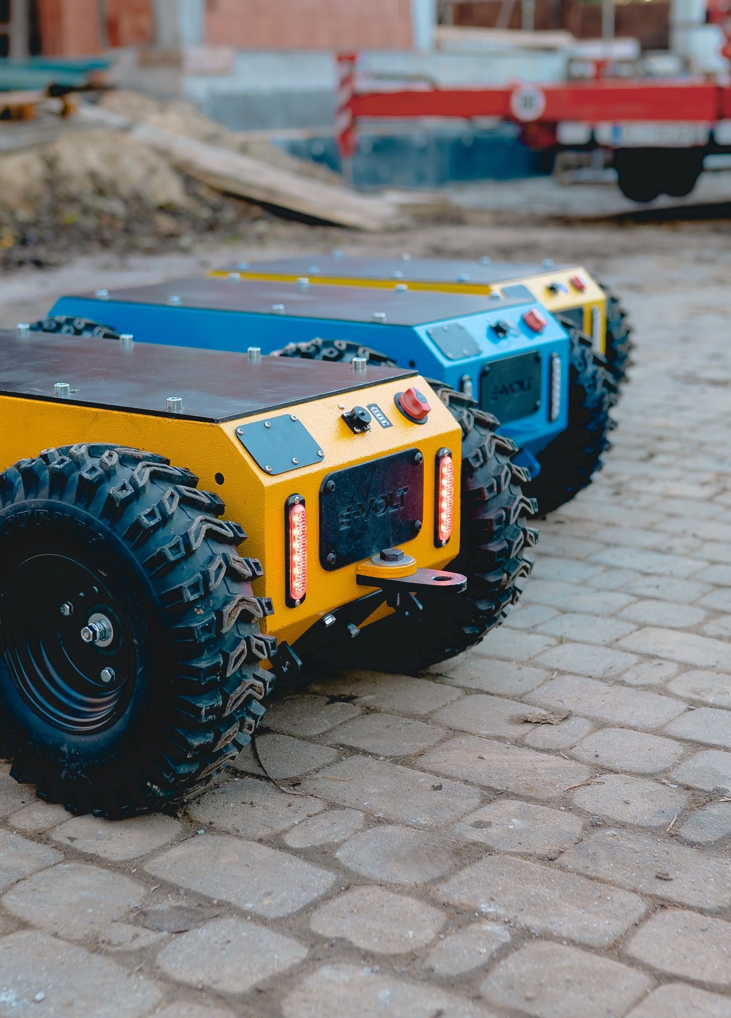Trzy metalowe platformy na kołach, w kolorach żółtym i niebieskim, z oświetleniem LED, ustawione rzędzie na brukowanej nawierzchni. W tle elementy budowlane.