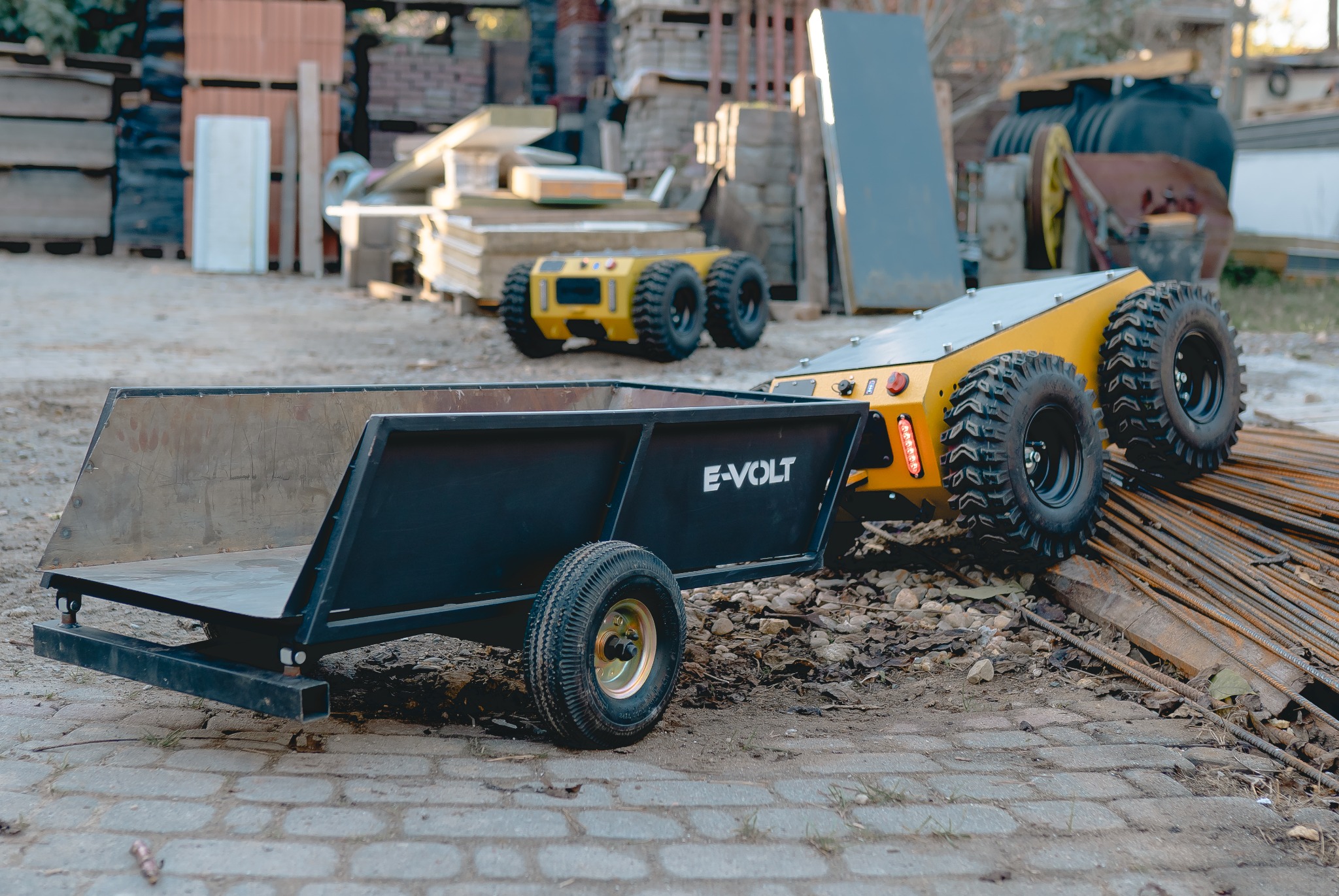 Żółty robot terenowy E-VOLT z czarną przyczepą na tle placu budowy. Widoczne materiały budowlane i drugi robot w oddali. Robot stoi na prętach zbrojeniowych.
