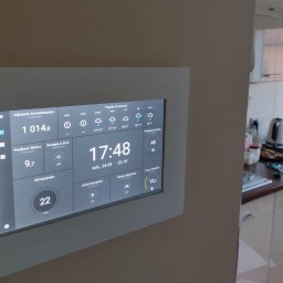 DEX-MAX PATRYK KOPERSKI - Panel sterowania inteligentnego domu z wyświetlanymi danymi pogodowymi, zużyciem energii i statusem urządzeń, wbudowany w ścianę w tle kuchni.