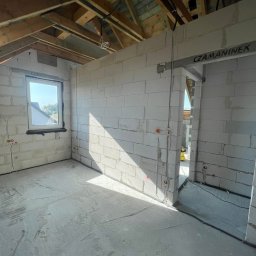 DEX-MAX PATRYK KOPERSKI - Surowy stan wnętrza z białych bloczków, widoczne wyprowadzone przewody elektryczne w ścianach i pod sufitem, drewniana konstrukcja dachu, wpadające światło słoneczne przez okno.