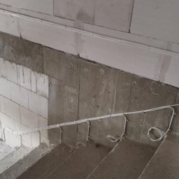 DEX-MAX PATRYK KOPERSKI - Surowe wnętrze w trakcie prac wykończeniowych: widoczne białe przewody elektryczne poprowadzone wzdłuż betonowych schodów i ściany z bloczków, z puszką elektryczną zamontowaną na górze.