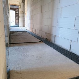 DEX-MAX PATRYK KOPERSKI - Wnętrze w trakcie budowy z widocznymi rurami instalacji elektrycznej ułożonymi na podłodze, ściany z białych bloczków, widok w głąb pomieszczenia przez otwarte drzwi.