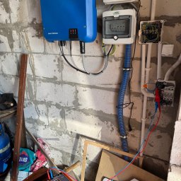 DEX-MAX PATRYK KOPERSKI - Szary bloczek betonowy w surowym stanie z zamontowaną niebieską skrzynką inwertera SMA Sunny Tripower, dwoma białymi skrzynkami rozdzielczymi Noark, oraz puszką elektryczną z przewodami;...