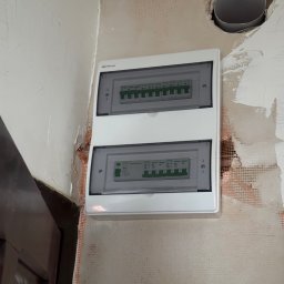 DEX-MAX PATRYK KOPERSKI - Biała skrzynka rozdzielcza z dwoma rzędami zabezpieczeń zamontowana na surowej ścianie z widoczną siatką i śladami po tynku, obok otworu w ścianie z wystającą rurą.