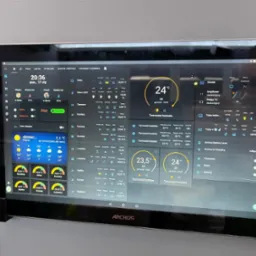 Tablet Archos wyświetlający interfejs inteligentnego domu z informacjami o temperaturze, alarmach i zużyciu energii w różnych pomieszczeniach, zamontowany na ścianie.