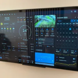 Ekran tabletu zamontowanego na ścianie prezentuje panel sterowania inteligentnym domem, z widocznymi informacjami o pogodzie, zużyciu wody, statusie alarmu, obrazem z kamery monitoringu...