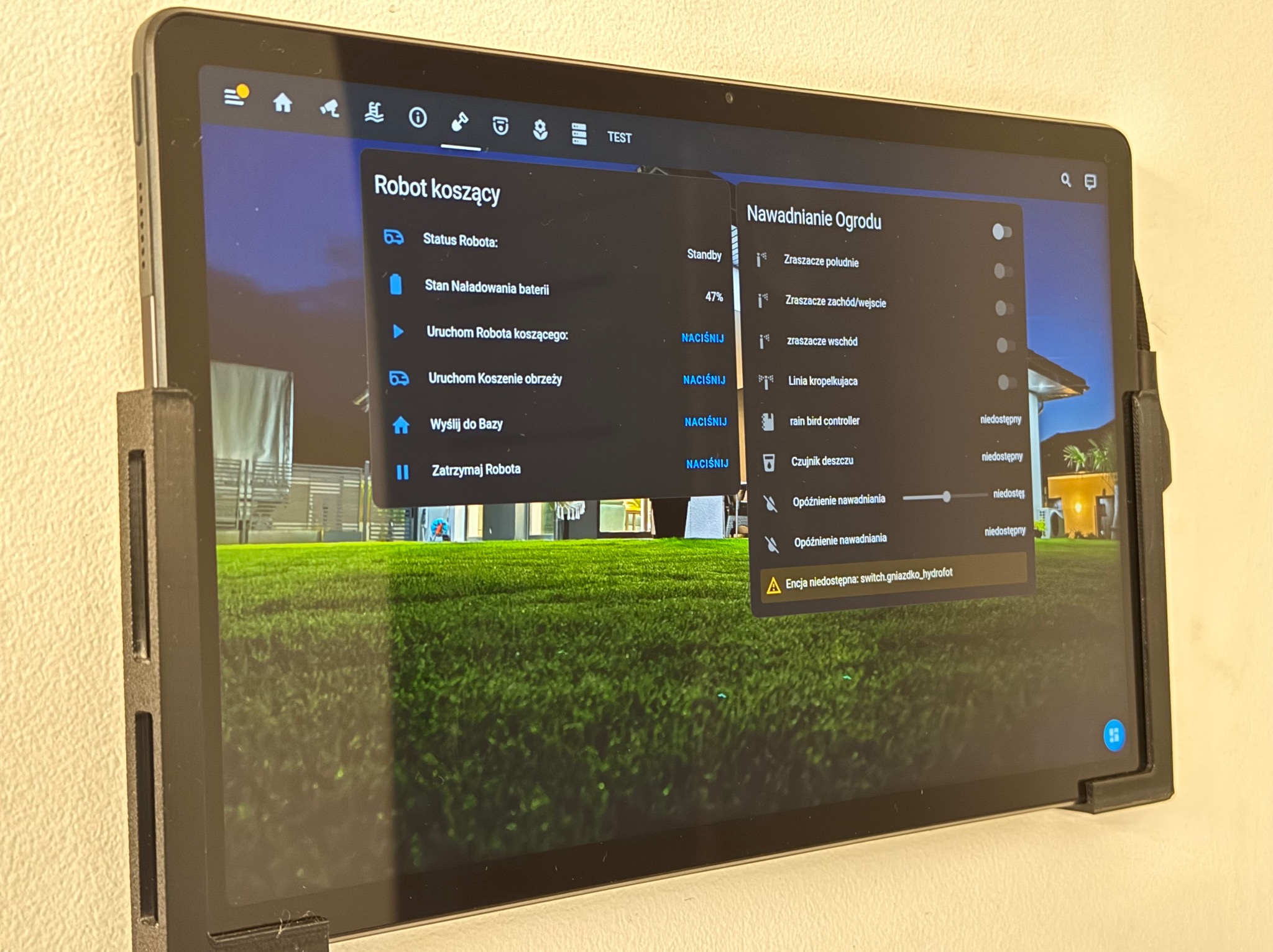 Tablet przymocowany do ściany, wyświetlający interfejs aplikacji do sterowania robotem koszącym i nawadnianiem ogrodu, z widocznymi opcjami konfiguracji zraszaczy i stanu baterii.