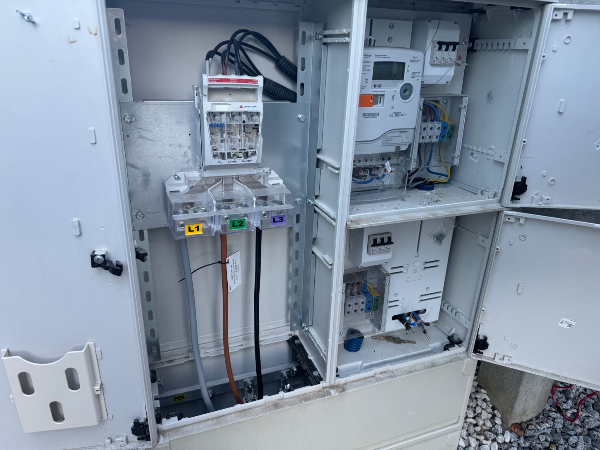 Otwarta skrzynka elektryczna z licznikiem, bezpiecznikami i okablowaniem, widoczne oznaczenia faz L1, L2, L3, oraz elementy montażowe na metalowej konstrukcji wewnątrz obudowy.