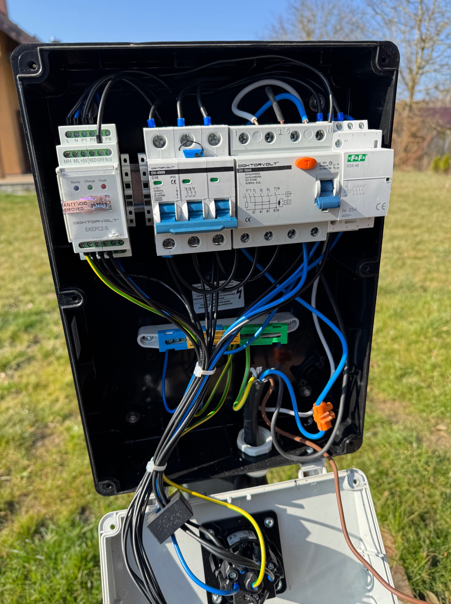 Otwarta skrzynka elektryczna z widocznymi bezpiecznikami, przewodami różnokolorowymi i modułem sterującym Doktovolt EKEPC2-S na tle trawnika w słoneczny dzień.