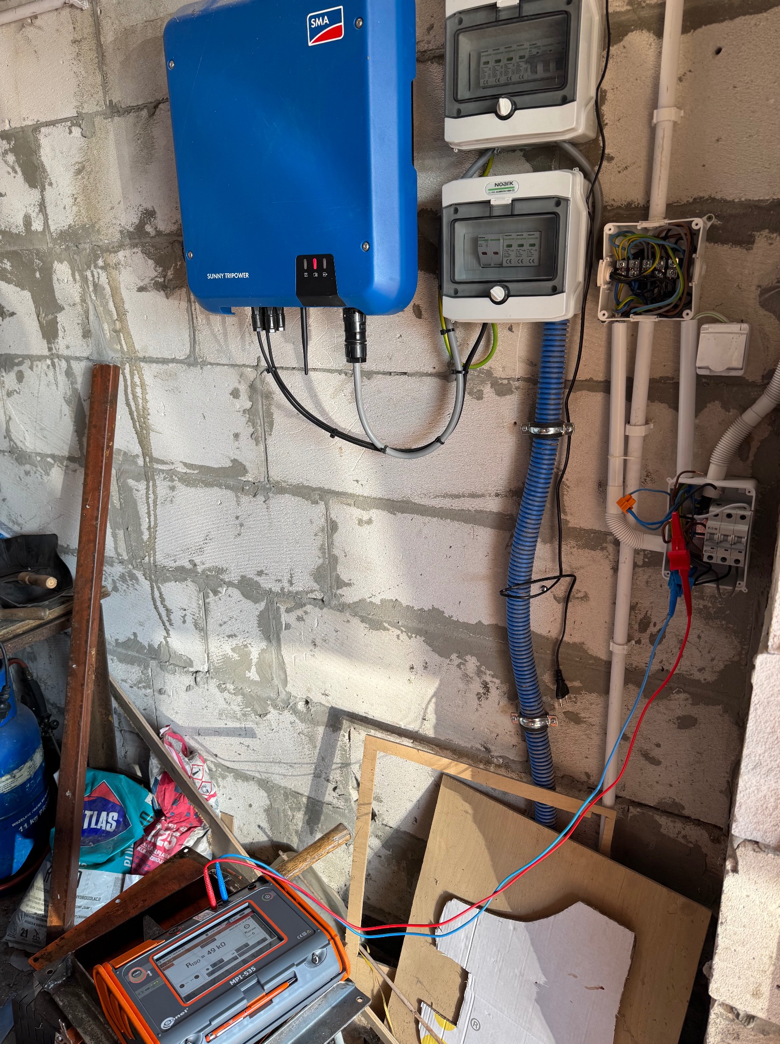 Szary bloczek betonowy w surowym stanie z zamontowaną niebieską skrzynką inwertera SMA Sunny Tripower, dwoma białymi skrzynkami rozdzielczymi Noark, oraz puszką elektryczną z przewodami;...