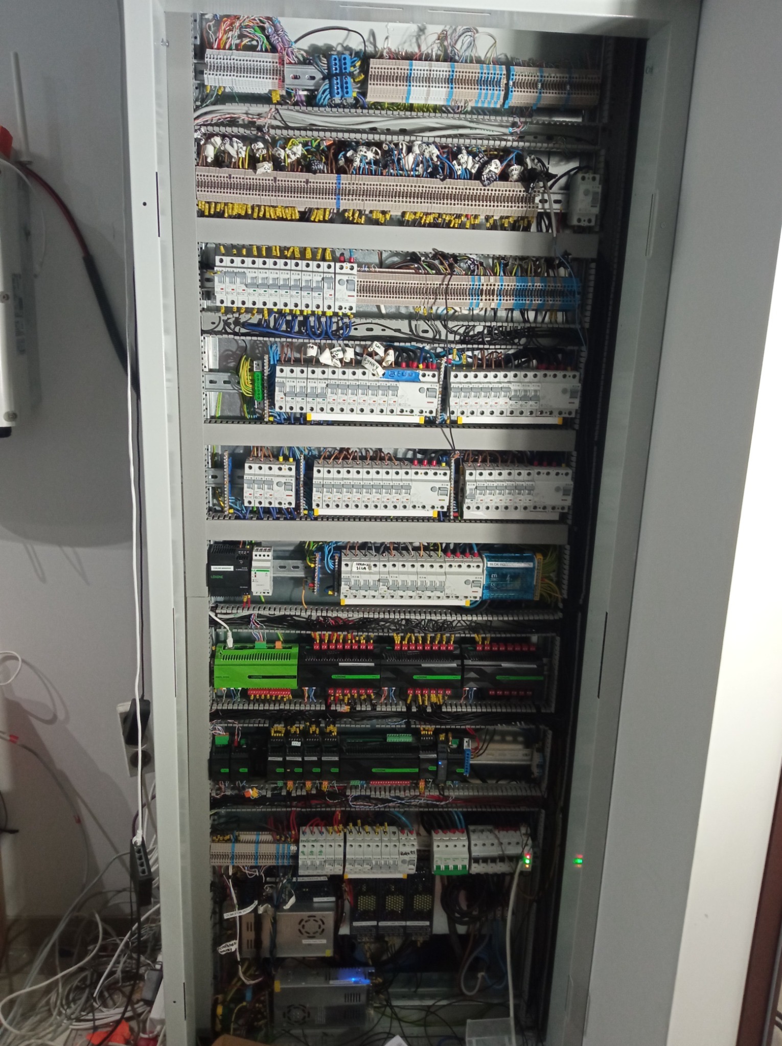 Otwarta szafa elektryczna z widocznymi licznymi przewodami, zabezpieczeniami, sterownikami PLC i listwami zaciskowymi, uporządkowana instalacja elektryczna w szarej obudowie.