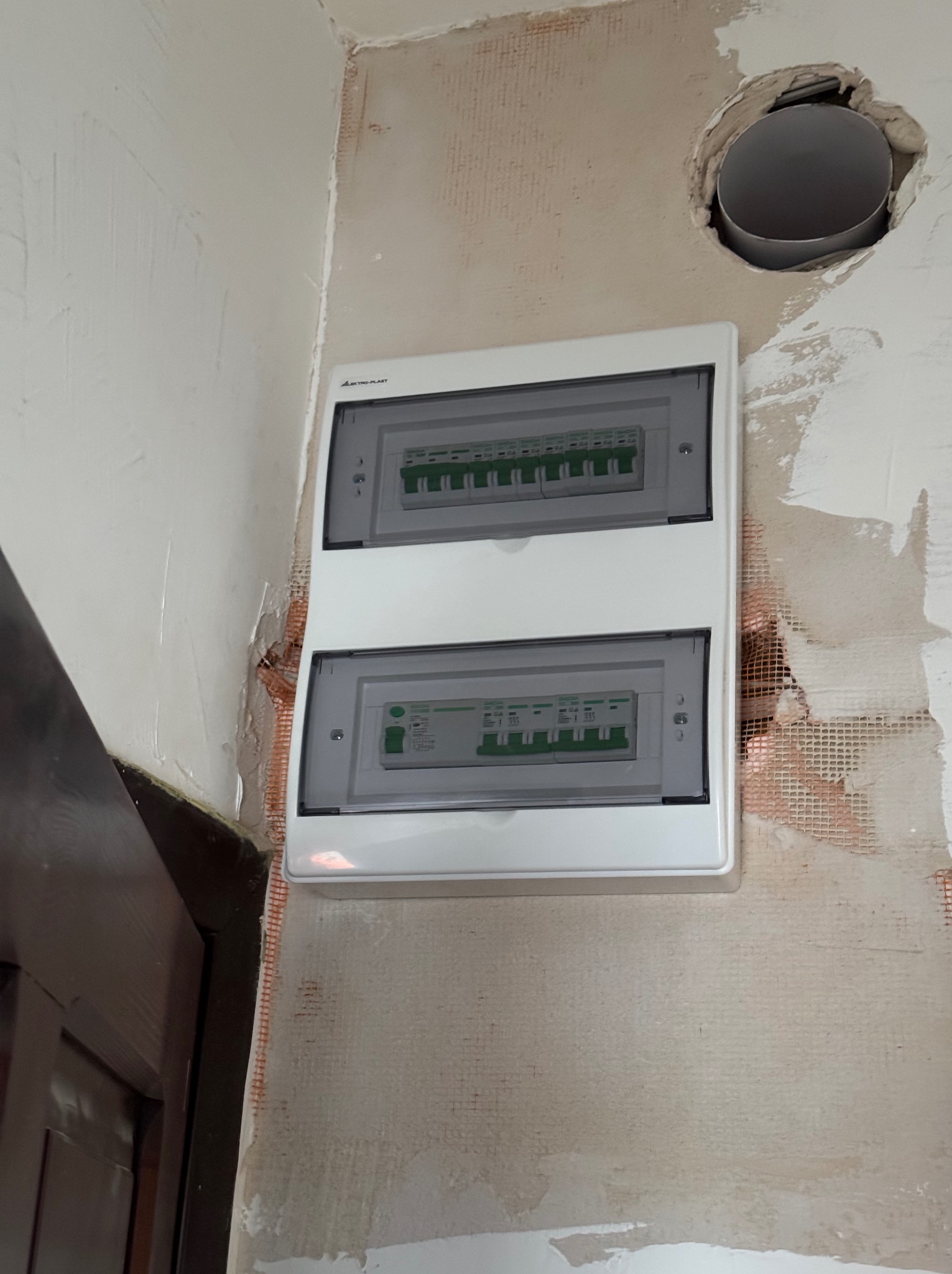 Biała skrzynka rozdzielcza z dwoma rzędami zabezpieczeń zamontowana na surowej ścianie z widoczną siatką i śladami po tynku, obok otworu w ścianie z wystającą rurą.