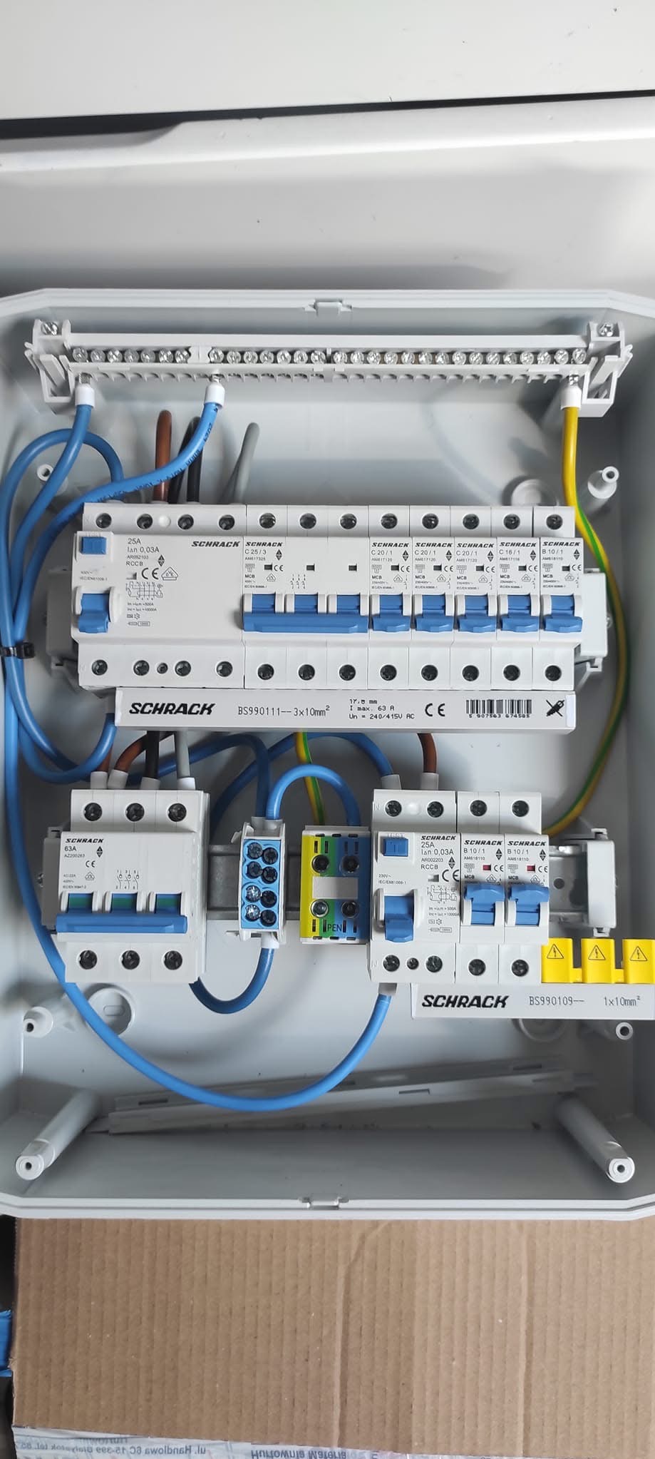 Otwarta skrzynka elektryczna z zamontowanymi bezpiecznikami Schrack, okablowaniem w kolorze niebieskim, brązowym i żółto-zielonym, widoczne złącza i oznaczenia modeli.