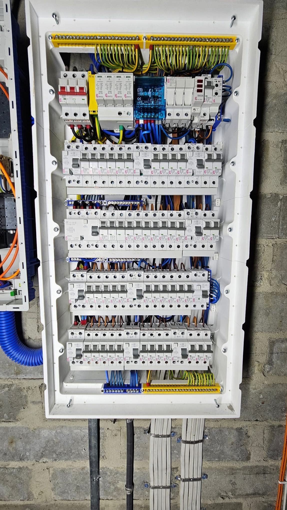 Rozdzielnica elektryczna z licznymi zabezpieczeniami, przewodami i listwami zaciskowymi w białej obudowie, zamontowana na szarej ścianie z betonu komórkowego. Widoczne kable doprowadzone do dolnej...