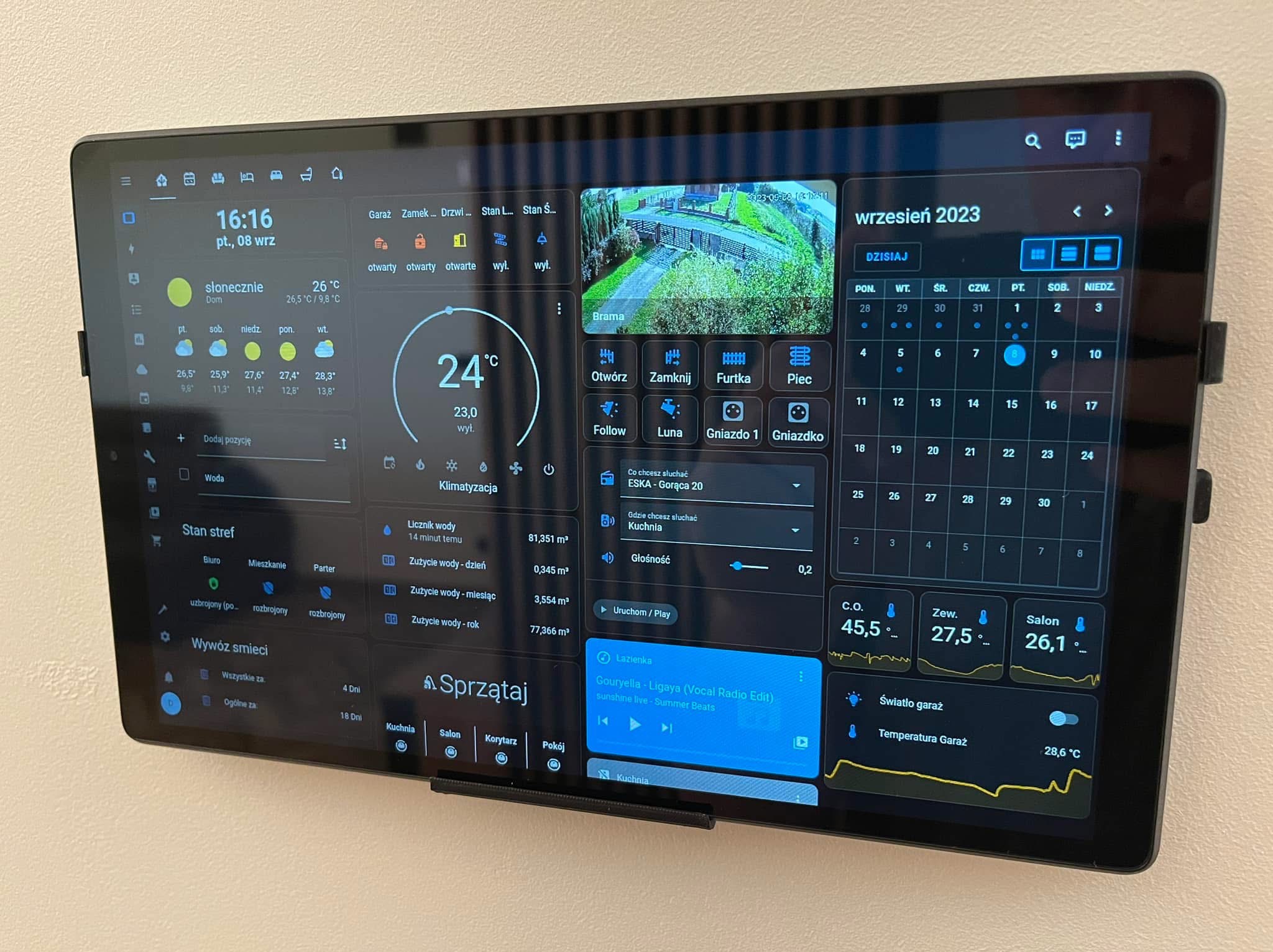 Ekran tabletu zamontowanego na ścianie prezentuje panel sterowania inteligentnym domem, z widocznymi informacjami o pogodzie, zużyciu wody, statusie alarmu, obrazem z kamery monitoringu...