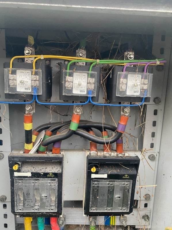 Rozdzielnica elektryczna z bezpiecznikami i przewodami oznaczonymi kolorowymi końcówkami, widoczne zabrudzenia i fragmenty roślin.