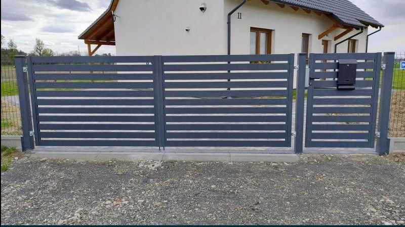Nowoczesna, grafitowa brama wjazdowa z poziomymi panelami i skrzynką na listy, widok z zewnątrz na tle białego domu z szarą dachówką i żwirowym podjazdem.