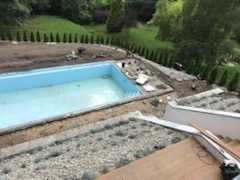 Widok z góry na basen w trakcie budowy, obok przygotowany teren pod kostkę, w tle iglaki i drzewa