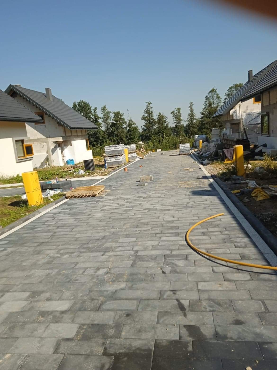 Ułożona, szara kostka brukowa na podjeździe między domami w trakcie budowy, widoczne żółte bariery i palety z kostką, błękitne niebo w tle.