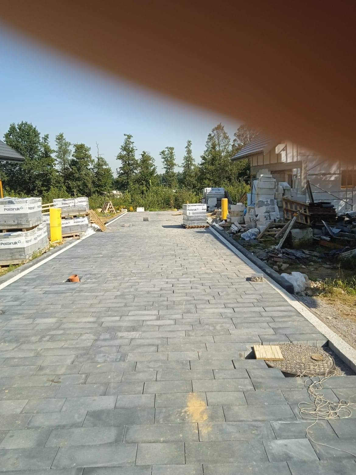 Ułożona szara nawierzchnia z kostki brukowej przed budynkiem w trakcie budowy, palety z kostką w tle, widoczne żółte słupki zabezpieczające i fragment instalacji kanalizacyjnej