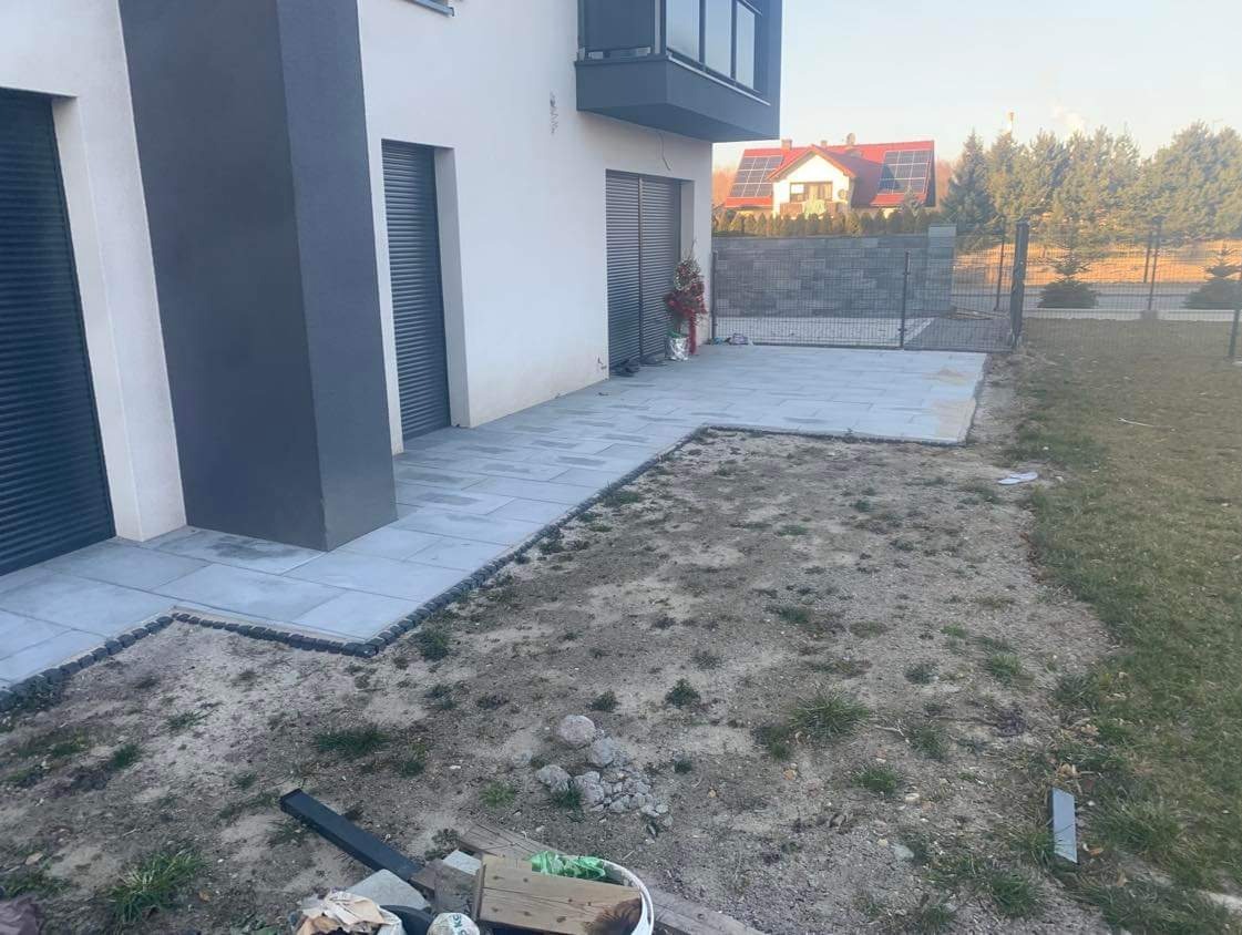 Nowoczesny taras i ścieżka z jasnoszarych płyt, obrzeża z ciemnych kamieni, widok na dom z szarymi roletami i ogrodzenie z siatki na tle drzew.