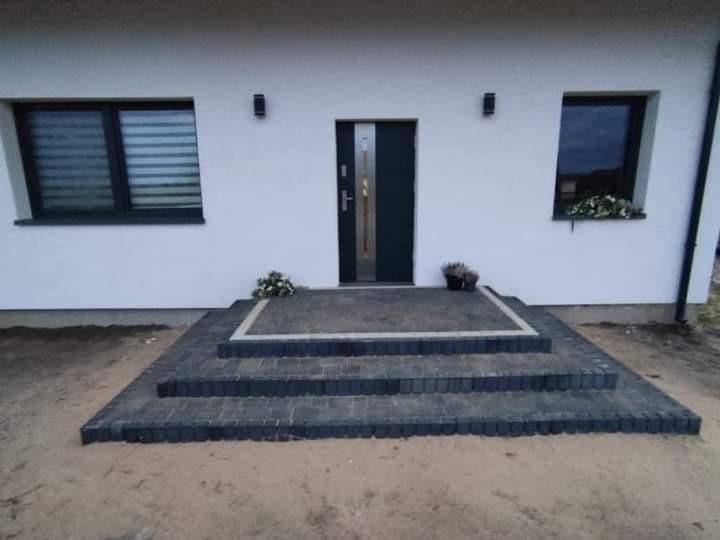 Schody wejściowe do domu wyłożone ciemnoszarą kostką brukową, z obramowaniem z jasnej kostki, kwiaty w doniczkach po bokach, elewacja budynku w tle.