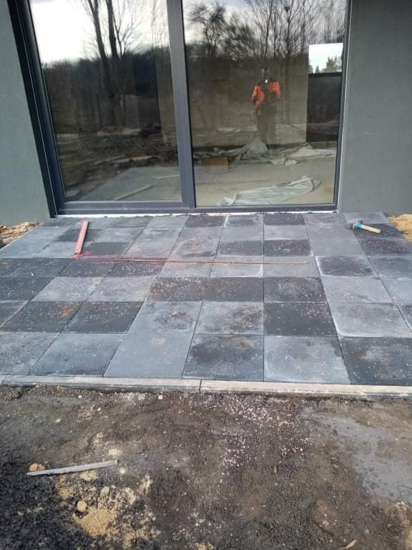 Układanie ciemnoszarej kostki brukowej w szachownicę na tarasie przy nowoczesnym domu z dużym oknem przesuwnym; poziomica i młotek widoczne jako narzędzia pracy.