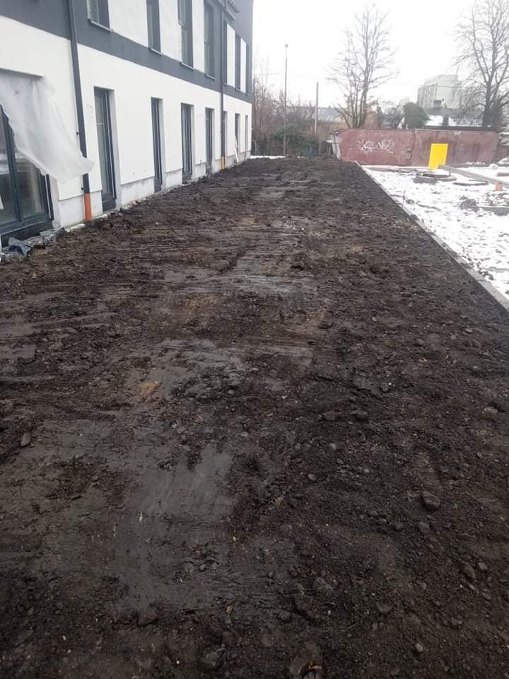 Przygotowany, wyrównany teren pod budynkiem, świeżo po niwelacji, z widocznymi śladami opon maszyn budowlanych, w tle fragment budynku i resztki śniegu.