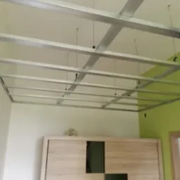 Metalowa konstrukcja sufitu podwieszanego z widocznymi elementami mocującymi, przygotowana do montażu płyt, z fragmentem zielonej ściany i szafy w tle.