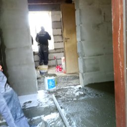 Grzegorz Zieliński Em-Bud Cieszyn - Wylewka podłogowa w trakcie realizacji. Widoczny pracownik, poziomica, wiadro z zaprawą. Surowy stan wnętrza z widocznymi elementami konstrukcyjnymi.