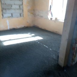 Grzegorz Zieliński Em-Bud Cieszyn - Wnętrze w trakcie prac: wylana, szara posadzka samopoziomująca w pomieszczeniu z oknem i ścianami w odcieniach beżu. Widoczny fragment kolumny.