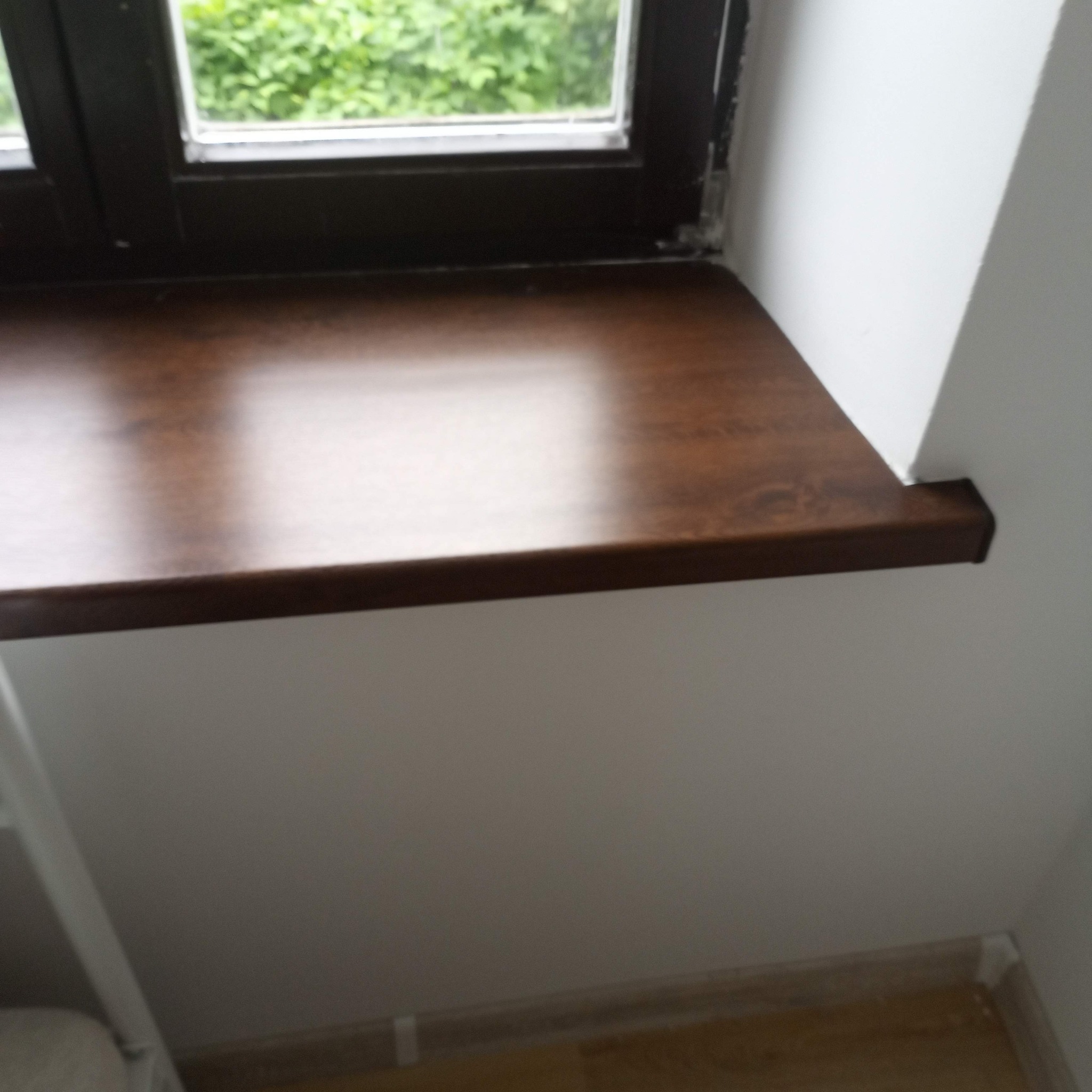 Błyszczący, brązowy parapet z drewna zamontowany pod oknem, widok z góry i boku, ściana pomalowana na biało.