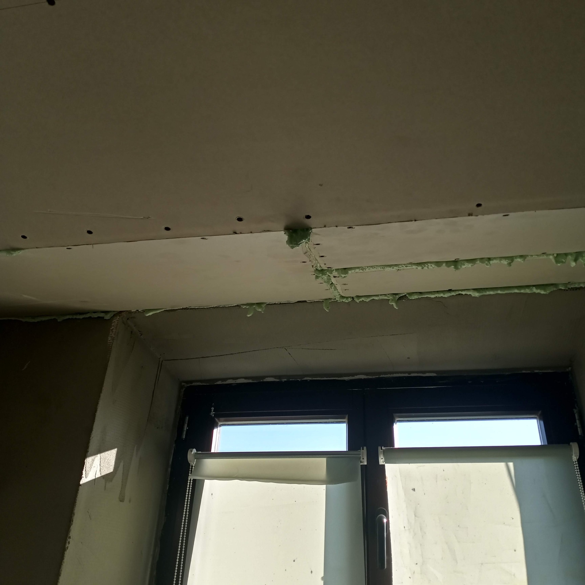 Montaż płyt karton gips na suficie w Cieszynie, widoczne wypełnienie pianką montażową między płytami, okno z roletami w tle.