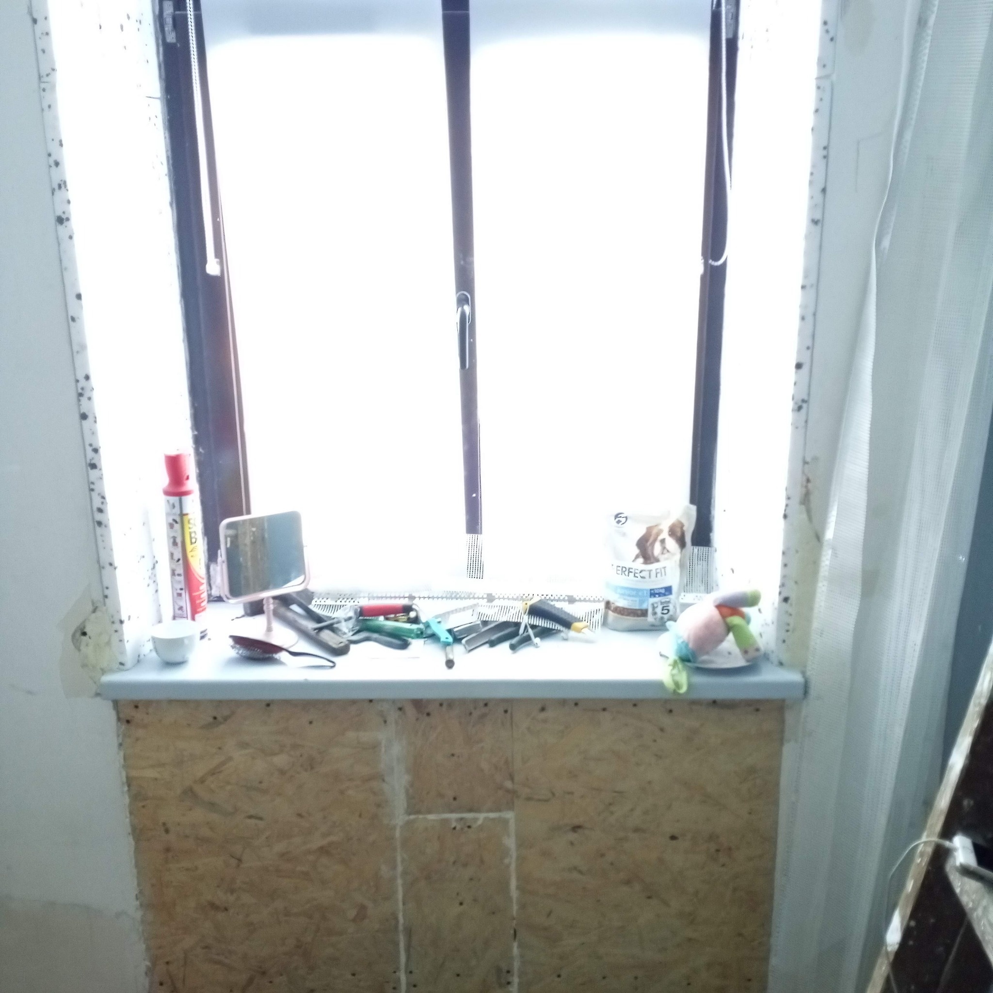 Przygotowany parapet z płyty OSB przed dalszymi pracami wykończeniowymi, widoczne narzędzia, puszka farby, lusterko i karma dla zwierząt na parapecie. Ściany w trakcie przygotowania do malowania.