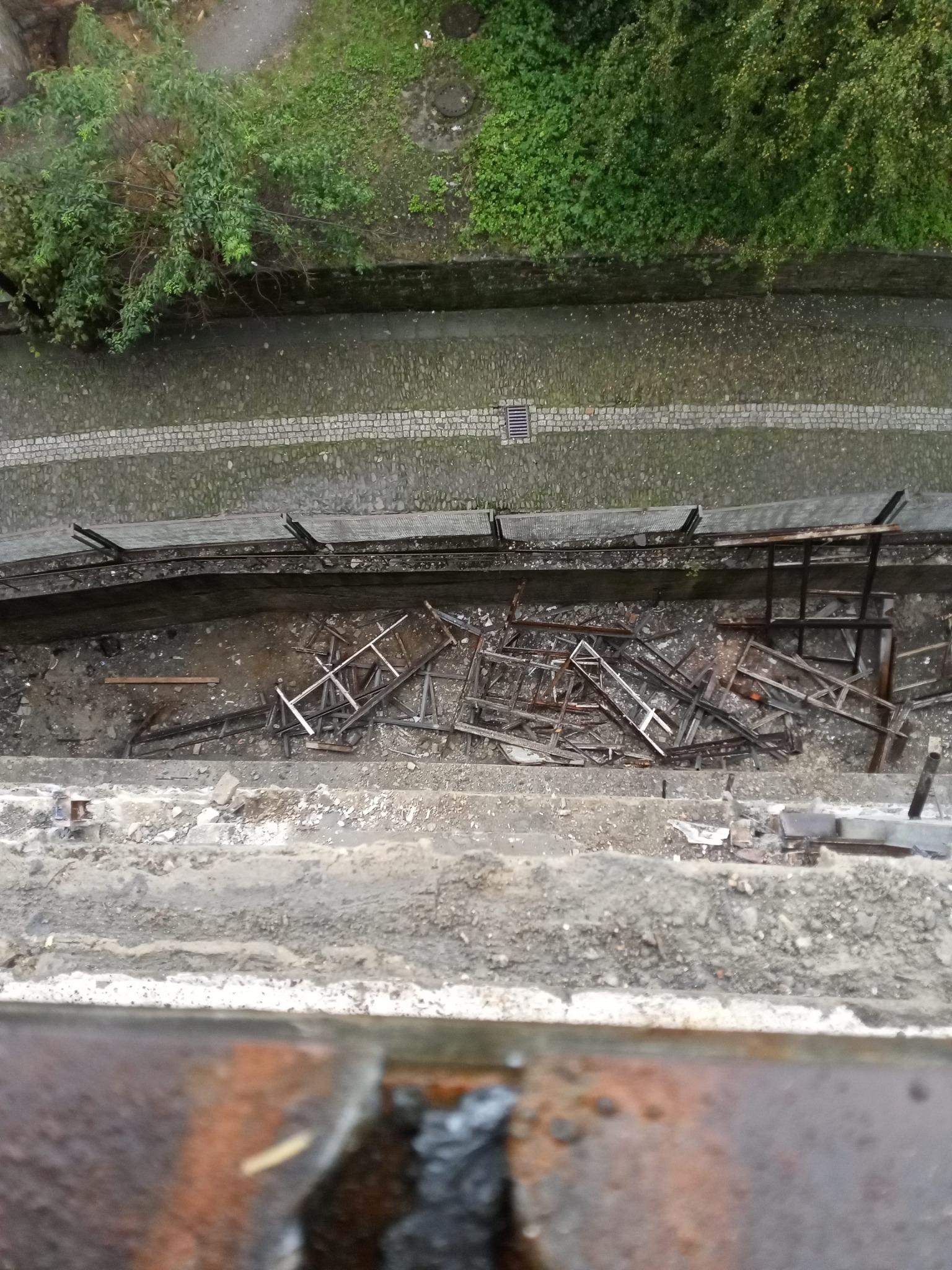 Widok z góry na plac budowy z rozbiórką konstrukcji drewnianej, gruzem i narzędziami, widoczny fragment chodnika z kostki brukowej i zieleni