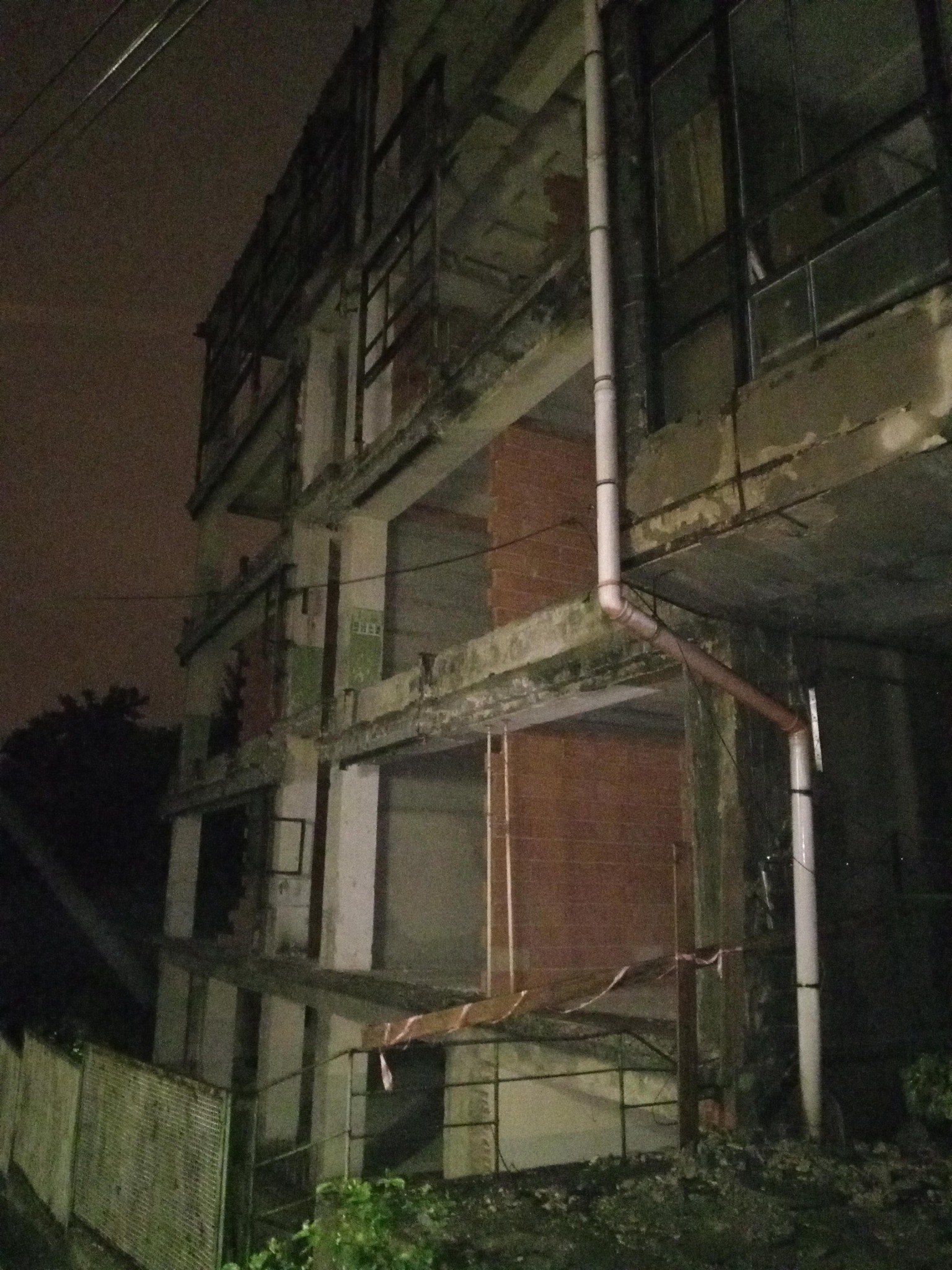 Niedokończony budynek w stanie surowym, widoczna cegła, rury spustowe, nocne oświetlenie, zabezpieczenia budowy.